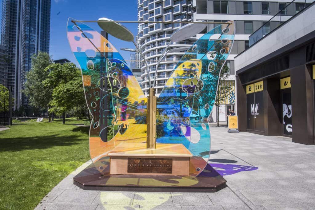 Out of the Cocoon in daylight on a plaza at Canary Wharf, its wings coloured blue, yellow and pink.
