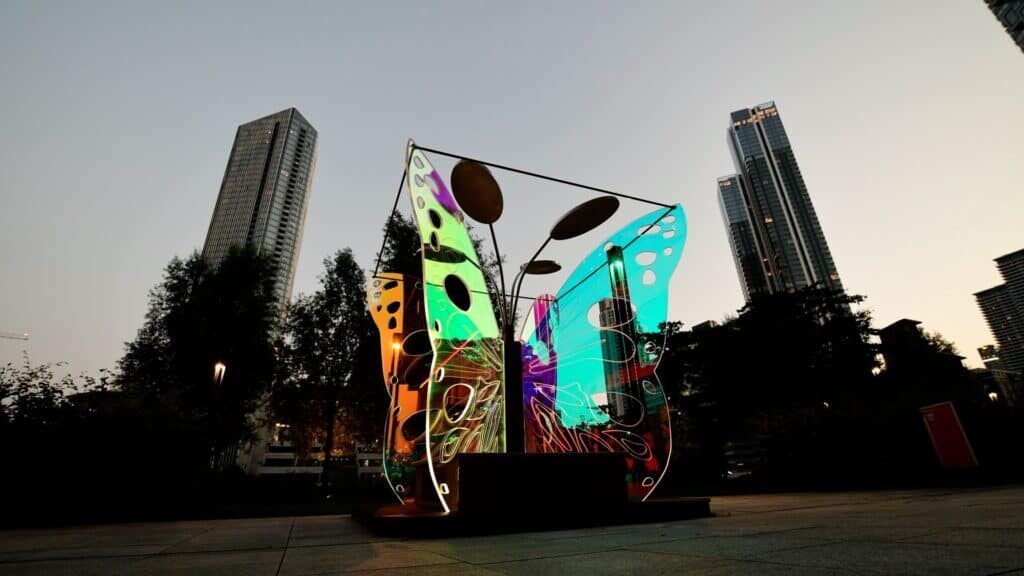 The Out of the Cocoon butterfly sculpture lit in green, blue and orange at dusk among the towers of Canary Wharf.