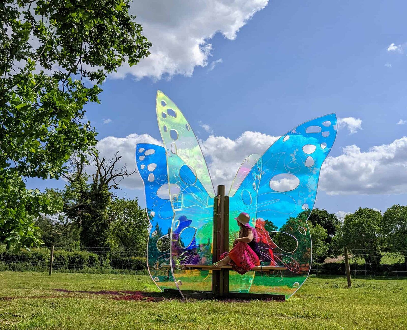 A visitor sitting beneath the iridescent wings of Out of the Cocoon in a green field under a blue sky.