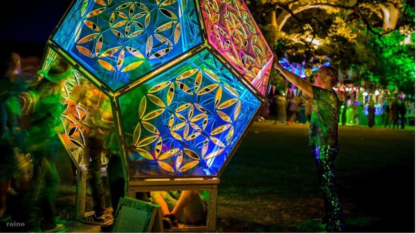 Seed of Life at a festival at night, its blue and green panels glowing, with visitors walking past.
