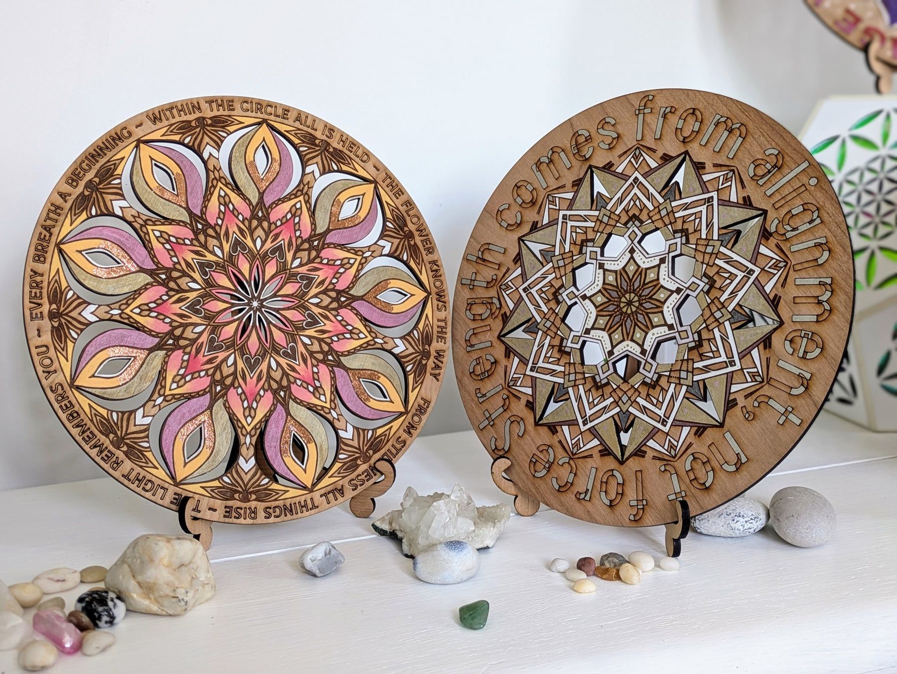 Two finished laser-cut wooden mandalas from Amber’s workshops, engraved with words and coloured in pink, gold and natural wood.